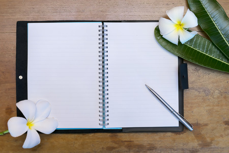 The notebook is open, white paper Have a line And with a stainless steel pen overlays, floral elements and leaves of the plumeria,The background is wooden floor.の写真素材
