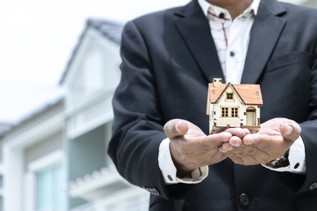 Businesses on buying - selling homes, businessmen with models, house put on hand, backdrop being home, wearing black suit, white shirtの写真素材