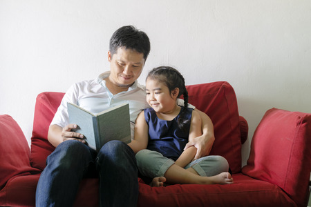 Father and daughter are reading books.Sitting on a red sofa.It is a good learning practice and a good family relationship.light shining.Like new things Good things are coming.の写真素材