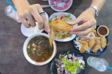 Thai food is very popular with people around the world. The hands of women who are catching fried chicken wings, Thai food and are eating it. The background has various types of food.の写真素材