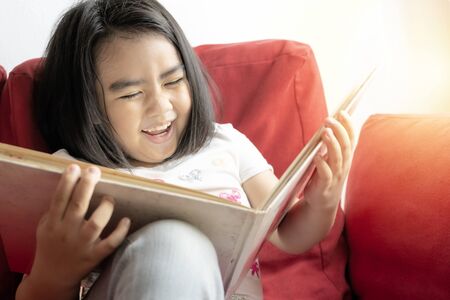 Cute asian girl She is very happy to be reading. Books can add imagination to her. Sitting on the red sofa happily.の写真素材