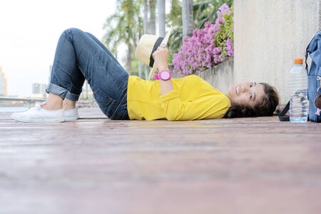 Women who are sleeping in the park happily. Wooden floor patio with space that can put text for advertising. Wearing a yellow shirt, jeans, white sneakers, a water bottle placed on the side.の写真素材