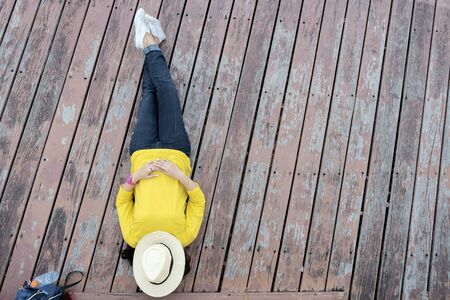 Women who are sleeping in the park happily on wooden floor patio. Wearing a yellow shirt, jeans, white sneakers, a water bottle placed on the side. She put a hat on the face to relax.の写真素材