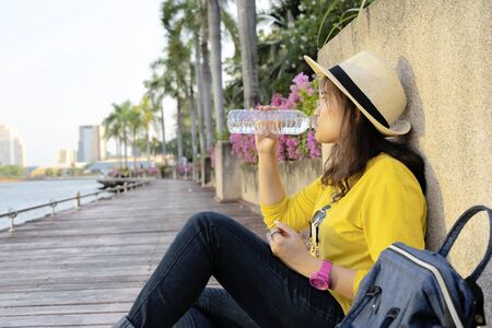 Beautiful young Asian tourists, yellow shirts, straw hats, backpacks Traveling in a park in Thailand. She was sitting and drinking pure clean water.の写真素材