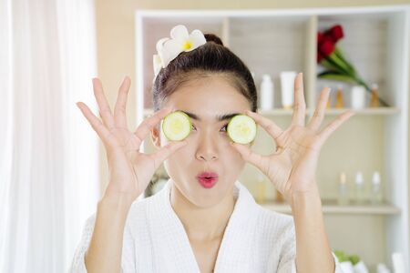 Young beautiful wearing a white bathrobes in a relaxed girl smiling hiding eye behind cucumber slice over spa room background. Beauty spa and cosmetology concept. casual atmosphere.の写真素材