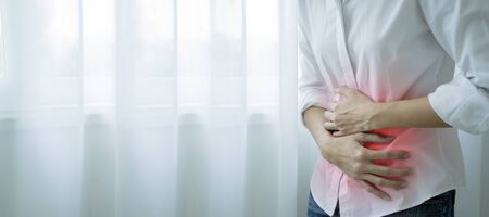 Upset stomach concept. Asian girl wearing white clothes standing in front of her window in the house with hand on stomach because nausea, painful disease feeling unwell. Ache concept, Panorama.の写真素材