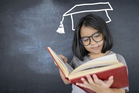 The cute girl wearing glasses is standing reading a book and she is congratulations graduates cap on blackboard, concept of education.の写真素材