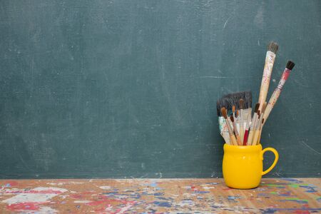 Many size paintbrushes are put in a yellow coffee mug. The floor has a lot of messy, blackboard background.の写真素材