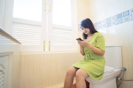 Cute Asian woman is sitting on the toilet, and is using a smartphone.の写真素材