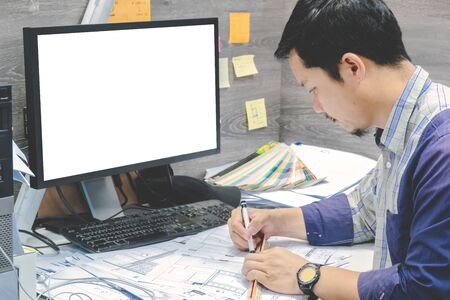 A young Asian architect sitting on a desk, designing the living room using a pencil, scale ruler Measured on the blueprint. The white computer monitor can insert text or mediaの写真素材