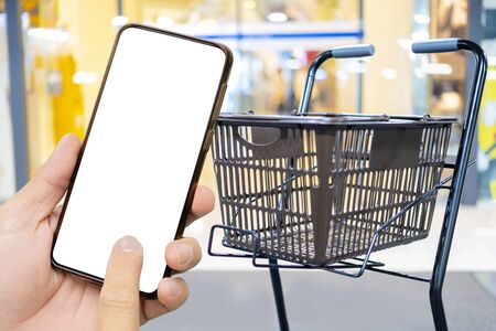 A man's hand holding a black smartphone, Touch screen smartphone in a hand. Shopping cart parked in front of the supermarket entrance, online shopping conceptの写真素材