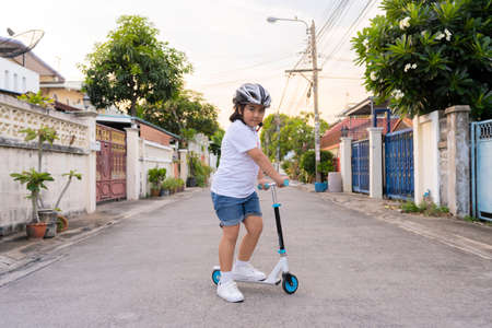 Young asian girl in safety helmet riding a roller. Kids play outdoors with scooters. Active leisure and outdoor sport for child.の写真素材