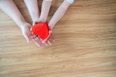 A red heart gift box in the hands of the child and mother on a white wooden background The concept...