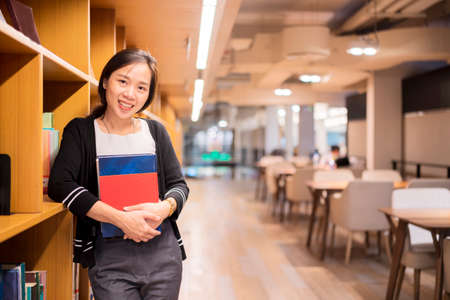 A cute Asian woman holding a book and smiling at the camera in the school library. University student conceptの写真素材