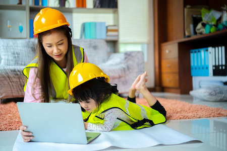 A beloved Asian mother and daughter pretend to be an engineer and smile with a safety hat in the house. Concept of builders and architects, using laptopの写真素材