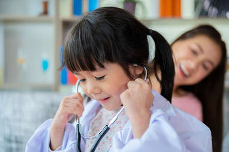 Cute little Asian girl act as nurse hold stethoscope listen to mom heart playing together at home, small Vietnamese child as doctor take care of mother or nanny, engaged in funny activity in bedroomの写真素材