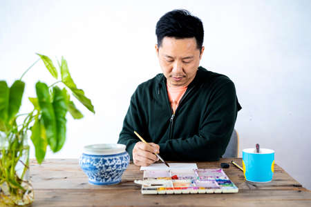 Asian male artist paints with a paint brush and watercolor palette on a laptop face paper.の写真素材