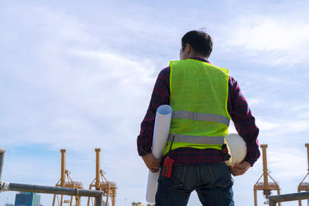 Engineer man wearing safety helmet and hold blueprints at a cargo port facility. Work outdoors Off-site exploration of the actual locationの写真素材