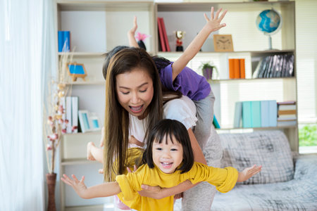 Happy family healthy cheerful young mum lifting cute little child daughter up playing plane doing gymnastic exercise in living room, funny kid girl flying in mothers arms look at camera having funの写真素材