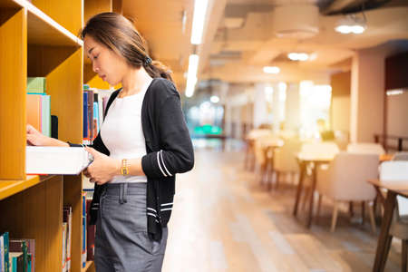 An asian woman standing in the library reading a book. Portrait of college girl reading book in libraryの写真素材