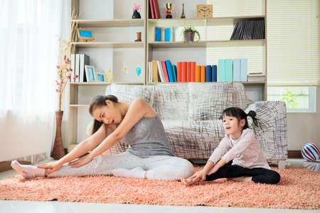 Beautiful young woman and her charming little daughter are smiling while doing yoga together at homeの写真素材