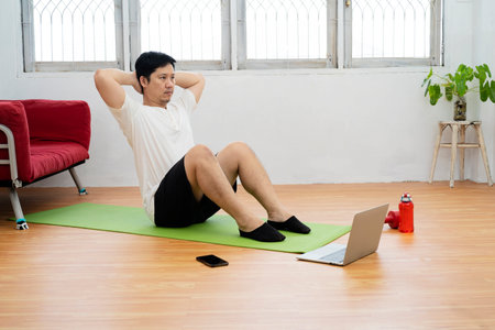 Handsome fitness man in a t-shirt and shorts doing abdominal exercises on floor at home. By searching for information from laptops and smartphonesの写真素材