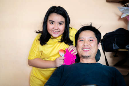 Funny little girl relax have fun with young father color paint lips with lipstick, happy smiling dad and preschooler daughter entertain enjoy spending time together do makeup relaxing on couchの写真素材
