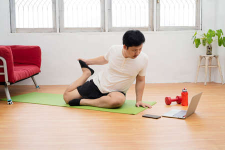 Active young cheerful man doing morning gymnastic at home, sitting on yoga mat and stretching, copy spaceの写真素材