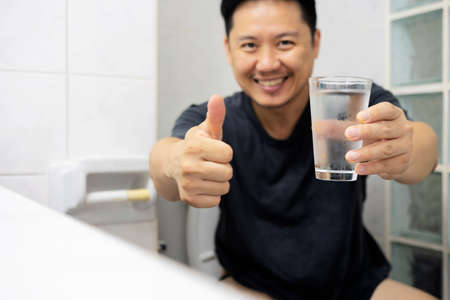 Close up satisfied man wearing t-shirt drinking pure clean mineral water in bathroom, enjoying...