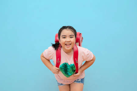 Happy school girl with bag and books posing at camera isolated over blue background, cheerful female...