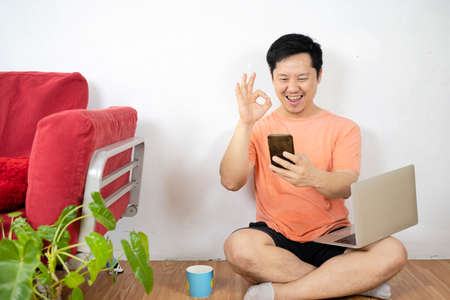 Smiling asian casual business man using zoom meeting online app, facetime video calling via mobile phone application, remotely working on laptop computer in coffee shop, social distancingの写真素材
