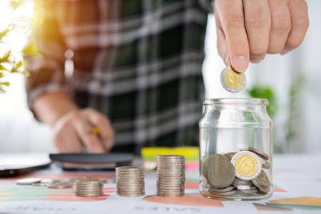 Closeup image of a businessman calculating, stacking and putting coins in a glass jar for saving money and financial conceptの写真素材