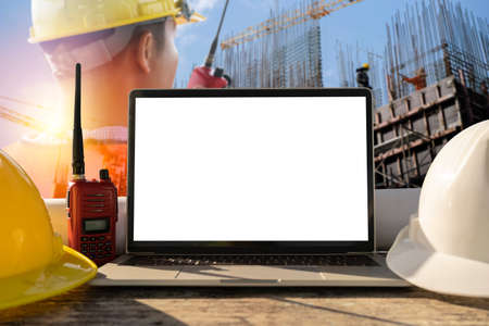 White and yellow safety helmet, laptops placed on a table in the construction area. Engineers are in control use communication tools, Heavy construction yellow crane for heavy lifting.の写真素材
