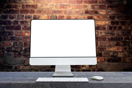 Computer monitor with white blank screen putting on wooden working desk with wireless mouse and keyboard over blurred vintage orange brick wallの写真素材