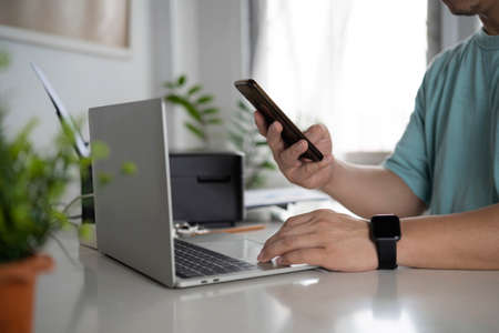 Business man using mobile smart phone, busy working on laptop computer browsing internet or checking internet application on smartphone with digital tablet and business data on office desk, close upの写真素材