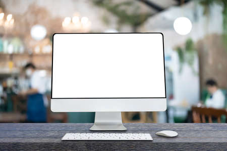 Computer monitor with white blank screen putting on wooden working desk with wireless mouse and keyboard over blurred restaurant as background.の写真素材