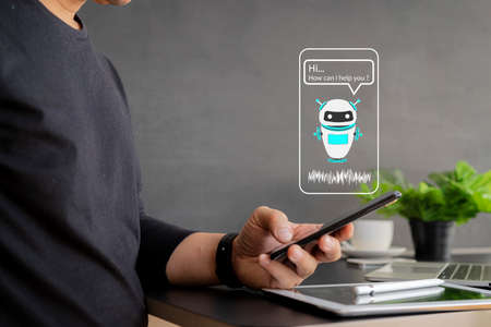 Chatbot assistant conversation, Ai Artificial Intelligence technology concept. Casual business man chatting with chatbot via mobile smart phone application while working on laptop computerの写真素材