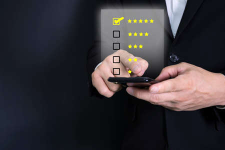 close up on customer man hand pressing on smartphone screen with gold five star rating feedback icon and press level excellent rank for giving best score point to review the service, business conceptの写真素材