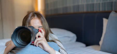 A beautiful woman in casual clothes is taking pictures with her camera while lying in bed at home. panoramic viewの写真素材