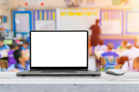Modern laptop with blank screen on table in classroom. Rear view of kids raising hands while teacher explaining the functioningの写真素材