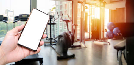 Hand holding smartphone device at gym background. interior of modern fitness center gym with equipmentの写真素材