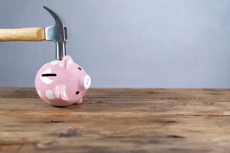 A hand holding a hammer which is raised above a pink china piggy bank, with a shocked and apprehensive facial expression. Reflective surface and light grey background. Copy space to the left.の写真素材