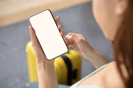 Back view of girl holding smartphone with white blank screen mockup. Traveler with phone at the airport with passport and suitcase. Lifestyle concept with digital technology.の写真素材