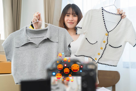 Asian cute female blogger online selling her clothes at home while using laptop and mobile phone to record video clip. Social media and technology concept.の写真素材