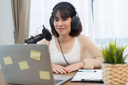 Creative smiling teenage blogger recording podcast audio in microphone sitting at table at home talking in microphone. Youth and blogging concept.の写真素材