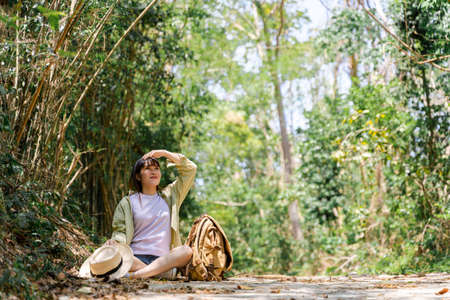 Cute young woman taking a break from forest walk in green tree background with straw fog, backpackの写真素材
