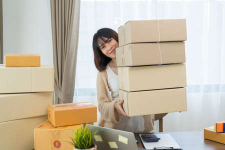 Happy young asian woman startup small business freelance holding parcel box and computer laptop and sitting on chair, Online marketing packing box delivery conceptの写真素材