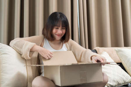 Smiling young woman unpacking parcel sitting on couch at home, pretty curious girl opening cardboard box close up, satisfied client received online store order, good delivery service conceptの写真素材