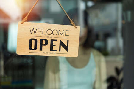 Store owner turning open sign broad through the door glass and ready to service. Open sign hanging front of cafe with colorful bokeh light abstract background. Business service and food concept.の写真素材