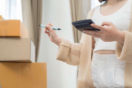 Young Asian female counting purchase order parcel before sending to customer from home, using a calculator to calculateの写真素材